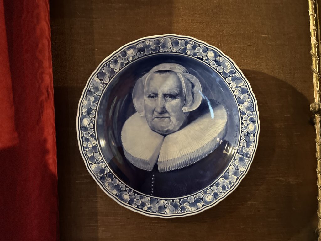An image of a plate hanging on a brown wall. The plate has a picture of a woman in a bonnet and a large ruffle collar painted in blue and white. Around the edge are florals painted in blue and white.