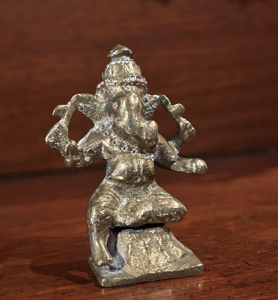 A small brass elephant. He has multiple arms and things in his hands and is sitting cross legged on a square stand.