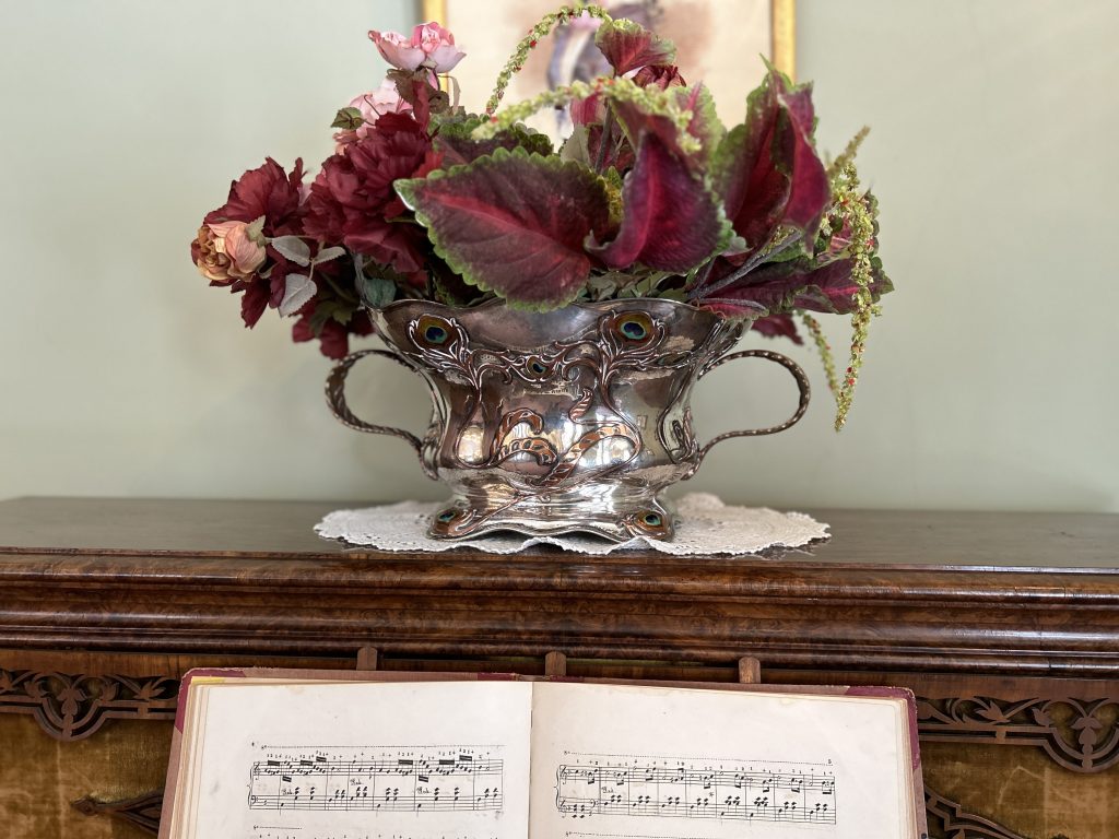 An image of a silver vase with two handles. The outside is decorated with enamel pieces that look like peacock feathers. Inside are fake red and orange flowers as well as coleus. It sits on a white doily and on a piano.
