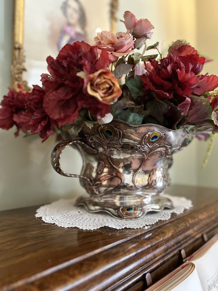 An image of a silver vase with large handles. It sits on an angle on top of a white doily and a wooden piano. Inside the vase there are fake red and orange flowers.