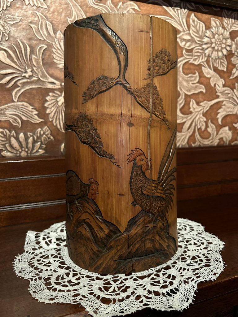 An image of a wooden cylinder. On it there are carvings of chickens and foliage. It is sitting on a white lace doily, in the background is orangish wallpaper.