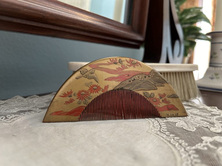 This is a Japanese hair comb that has a depiction of Japanese-style nature scenery with red flowers and a silver bird. It is placed on a white doily.
