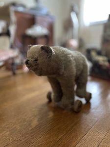 An image of a beige stuffed bear. He has wheels attached to his feet. He is sitting on a wooden floor, the background is blurred.