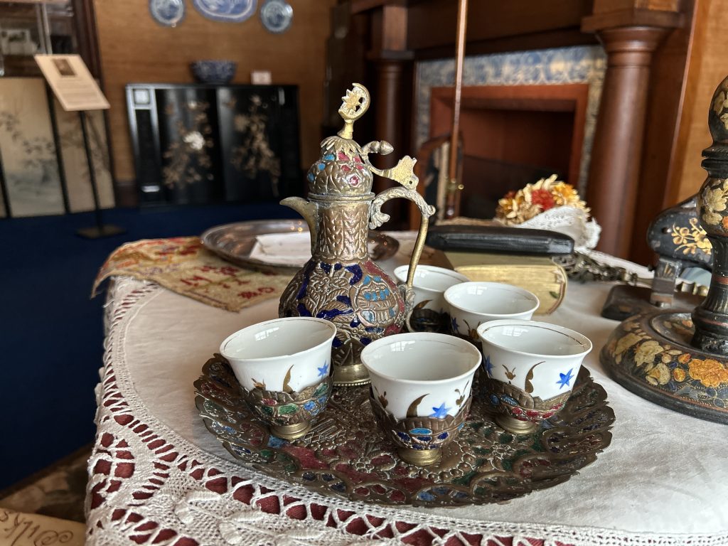 This is an ornate brass Turkish coffee set with blue, green, and red enamelwork. It is sitting on top of a matching brass serving tray on a white table cloth.