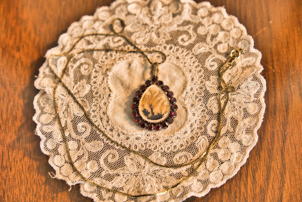 A tear drop shaped necklace with a willow tree on the inside. The willow tree is made of hair. This necklace is sitting on a white lace doily on top of a wooden table.