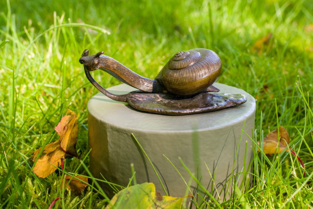An image of a metal snail on a leaf sitting on a round grey box. The box is sitting on grass amongst green leaves.