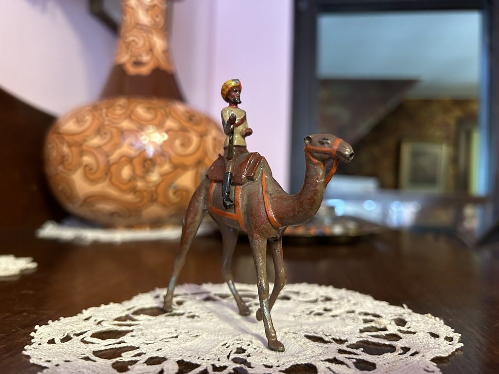 An image of a figurine of a man on a camel. He is sitting on a doily, and in the background is a orange vase.