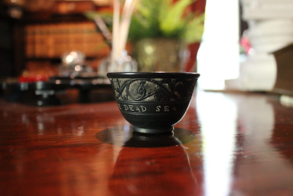 An image of a black stone bowl on a rich brown wood table. The bowl has light etching at the top. Under the words Dead Sea are visible.