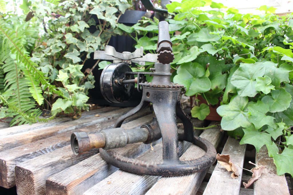 A metal sprinkler sitting on top of a slated wooden table with greenery behind.