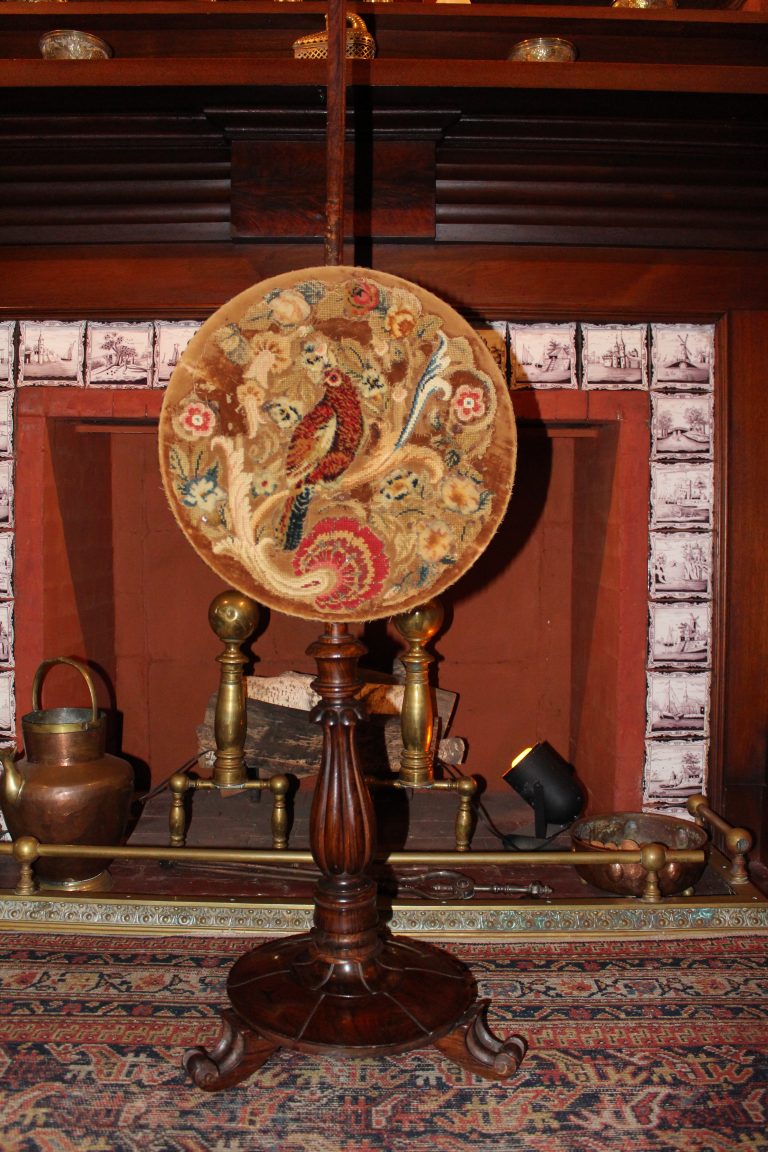 A large wooden pole sitting in front of a fireplace with purple and white tiles. The circular part of the screen is embroidered with birds and flora.