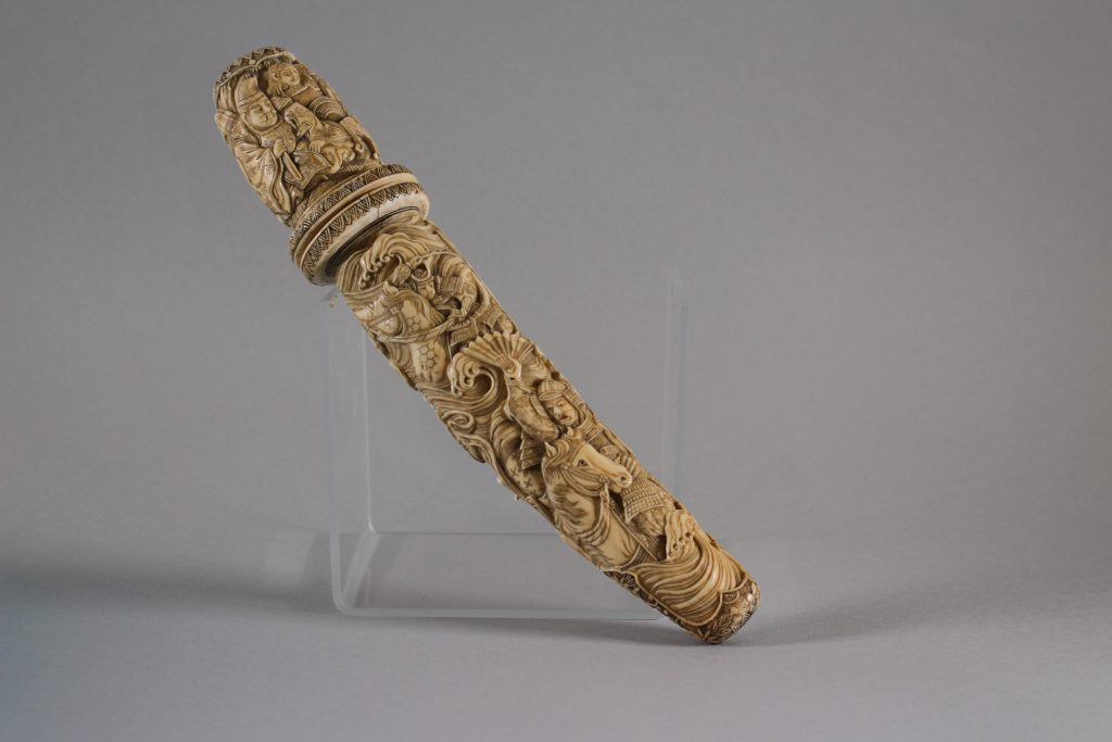 An image of a sheath against a grey background. The sheath has carved people, horses, and dragons on it.