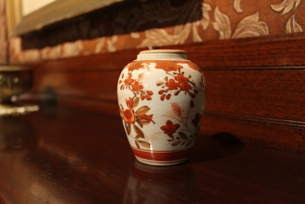 A small white jar decorated with orange brown and hints of gold in floral patterns. The jar is sitting on a dark wood shelf.