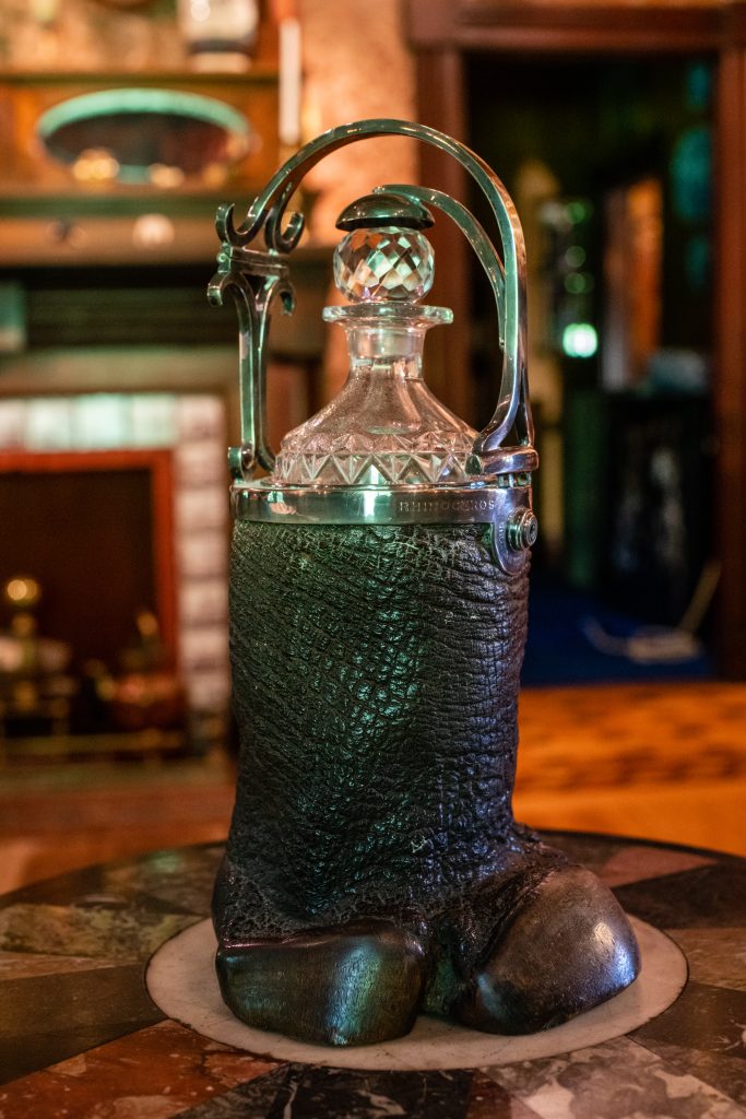 An image of a rhino foot with a metal rim on the top edge. Inside of the foot is a crystal bottle. It is sitting on a marble table.