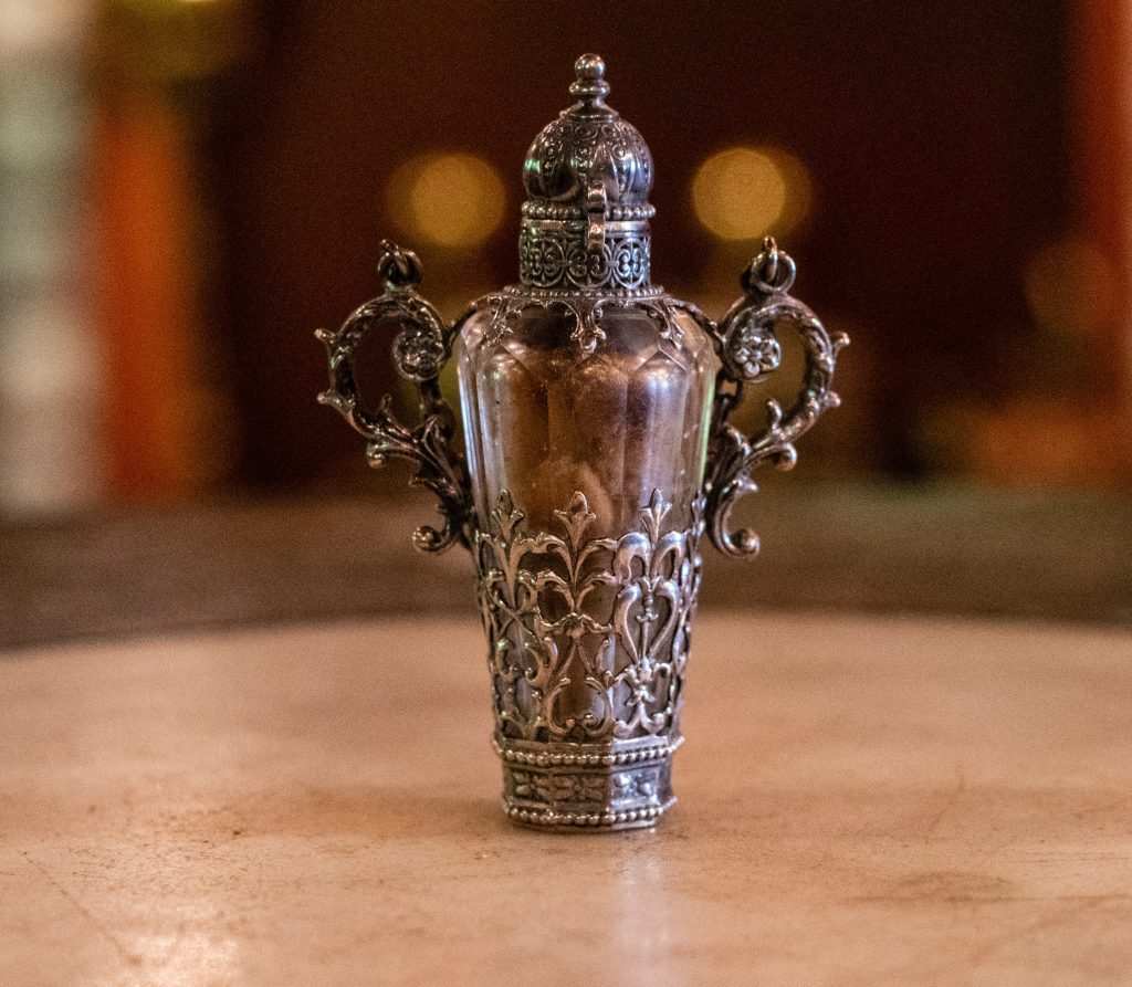 An intricate silver bottle with swirl handles on either side and scrollwork on the bottom. The bottle is sitting on an ivory table.