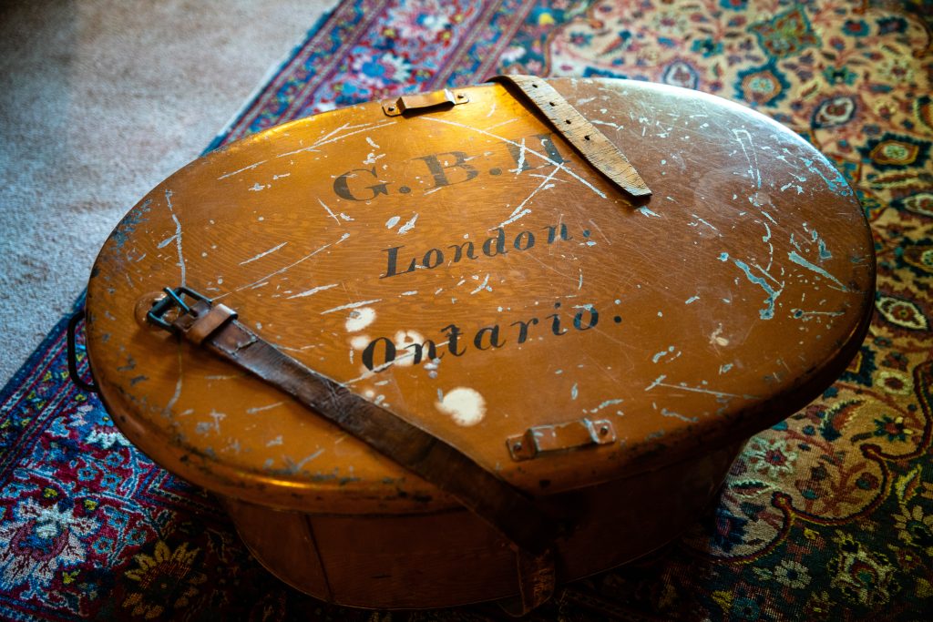 An image of an oval tan item. Straps come up to the worn lid. On the lid it says G.B.H. London, Ontario. It is sitting on a heavily patterned carpet