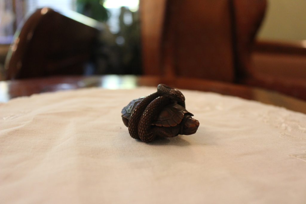 A small figurine of a turtle. A snake has coiled itself around the center of the turtle. The netsuke is on a white fabric doily on a wood table.