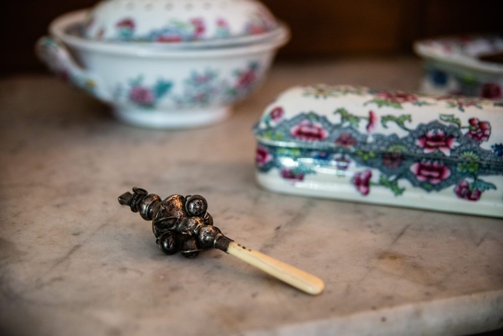 A small silver rattle with bells made of silver on top. The handle is made of a yellowish ivory. It is sitting on a marble table and in the background there are pots with pink flowers.