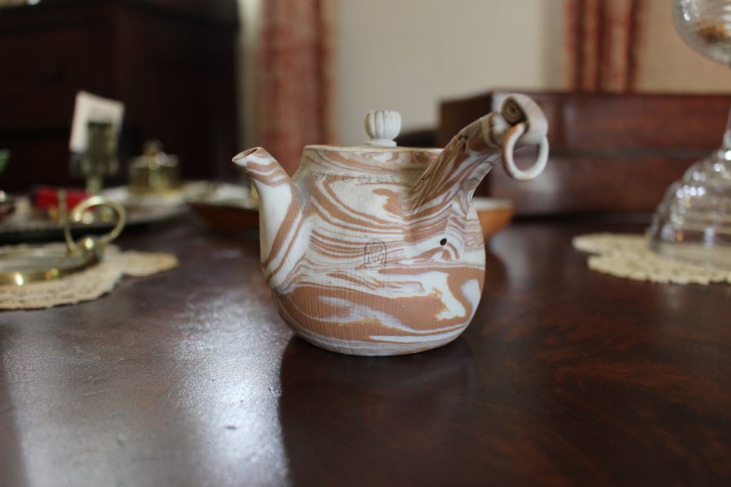 A small teapot with a large handle. Marbled white and brown. It is sitting on a brown table.