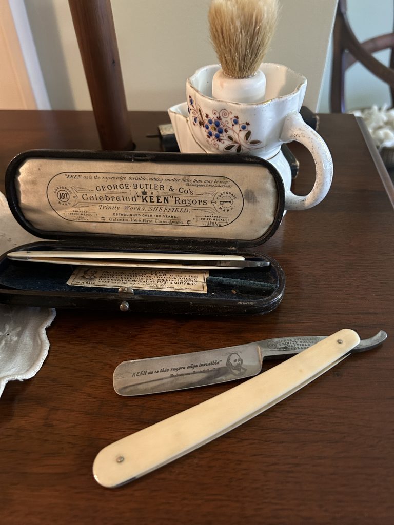 An image of a old school straight blade with a ivory handle. On the blade there is a quote and an image of William Shakespeare. In the background there is an open case with another razor blade. The case has an insert advertising for the razor company, Keen's. In the background there is the shaving brush and cup.