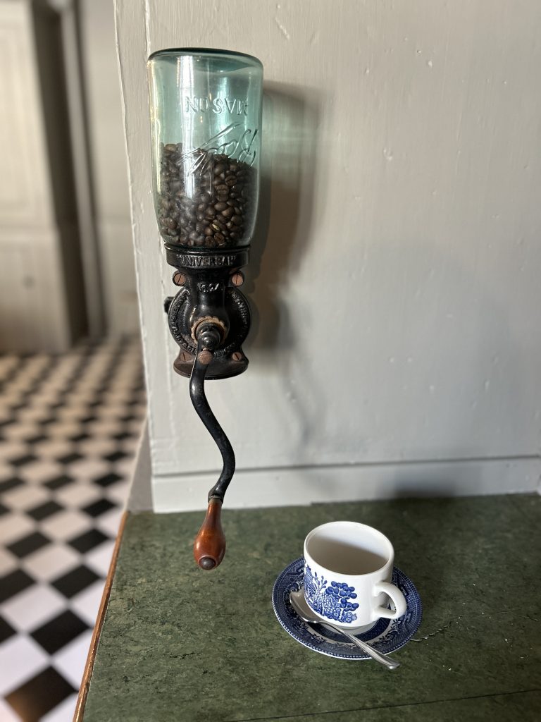 A large blue glass coffee grinder with beans in it. It is mounted on a grey wall and a green table. A coffee cup sits in front.