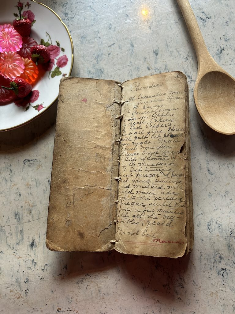 A worn thin paper book. A hand written recipe for chowder is on the right side. In the background is a wooden spoon on the right and a rose printed plate with pink jelly on the left.
