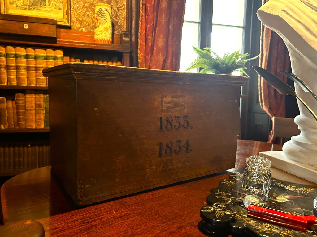 A large wooden box. On the front it says 1833. and 1834. In the background there are books and in the foreground writing tables.