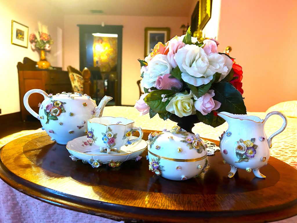 An image of bright white china tea set including a pot, cup and saucer, sugar bowl with a lid, and a creamer. All have brightly coloured roses and leaves stuck on the outsides. It is on a small wooden table with a vase of pink and white roses behind it.