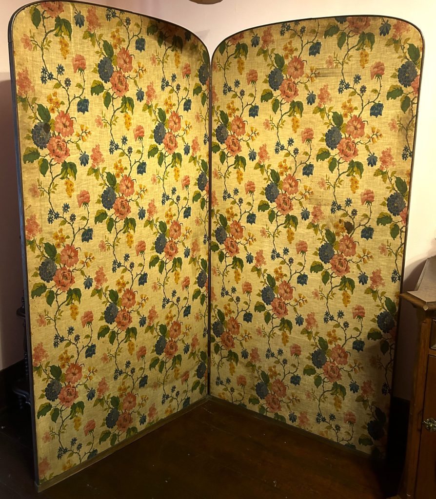 An image of a large yellow fabric folding screen. On the screen are pink and blue flowers with smaller yellow flowers, and green vines and leaves.