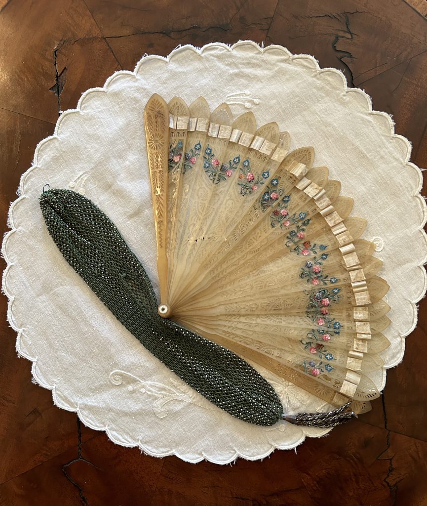 An image of a ivory fan with pink and blue flowers on the top. Under is a long green sparkly fabric piece, which is the fan holder. It is sitting on a white doily on top of a wood table.