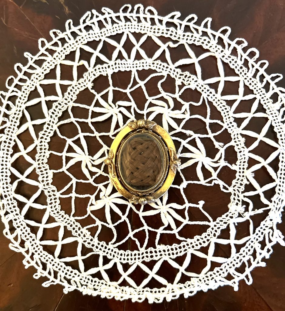 An image of a gold framed pendant. Inside there is braided light brown hair. It is topped with glass. Under there is a netted white doily sitting on a wood table.