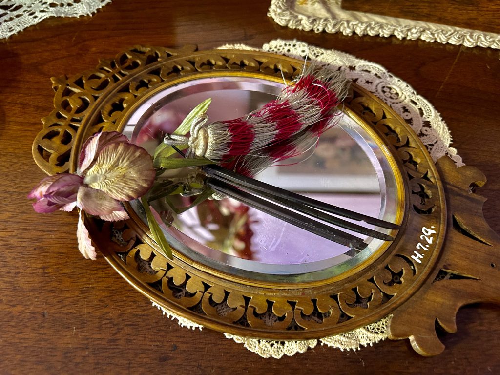 A wooden mirror with an ornate frame. Sitting on it the glass is a hairpin that has spikes. There is a pink flower and has a tassel.