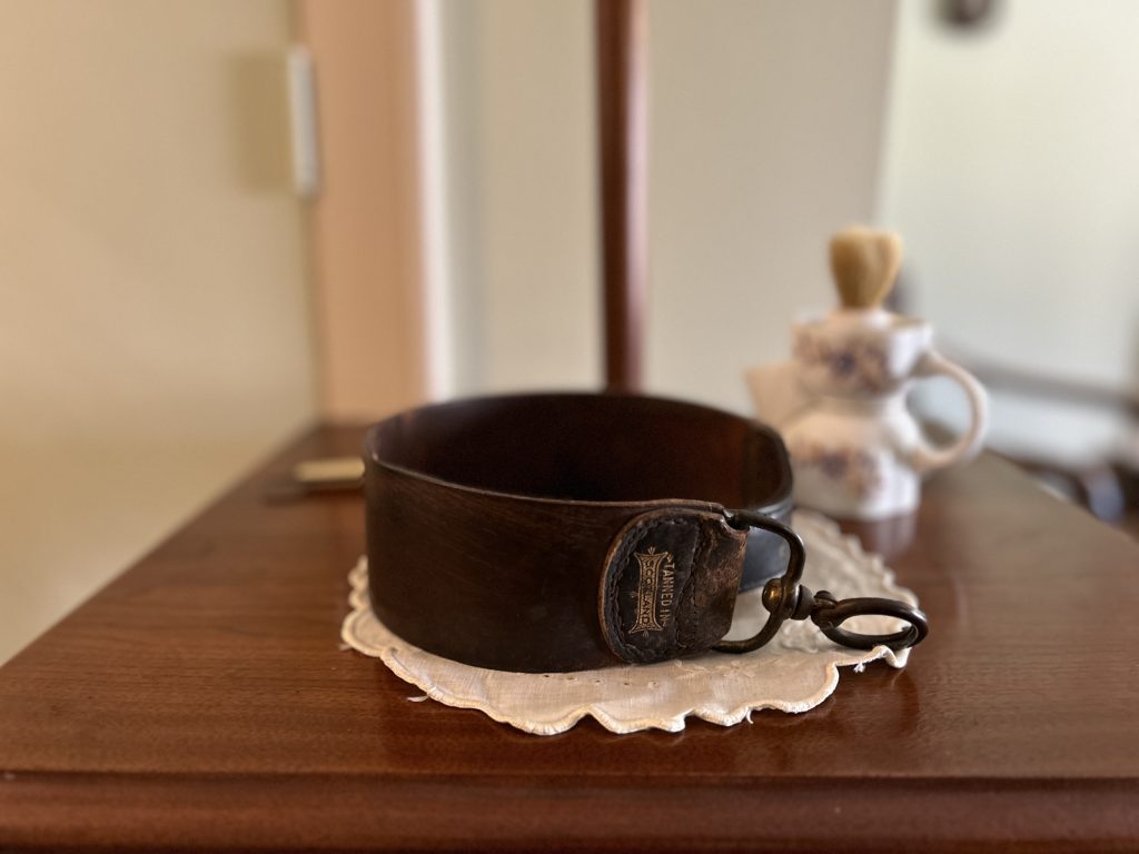 An image of a leather piece with a metal hanging portion at the end. In gold it says Tanned in Scotland. It is sitting on a white doily. In the background there is a white porcelain shaver.
