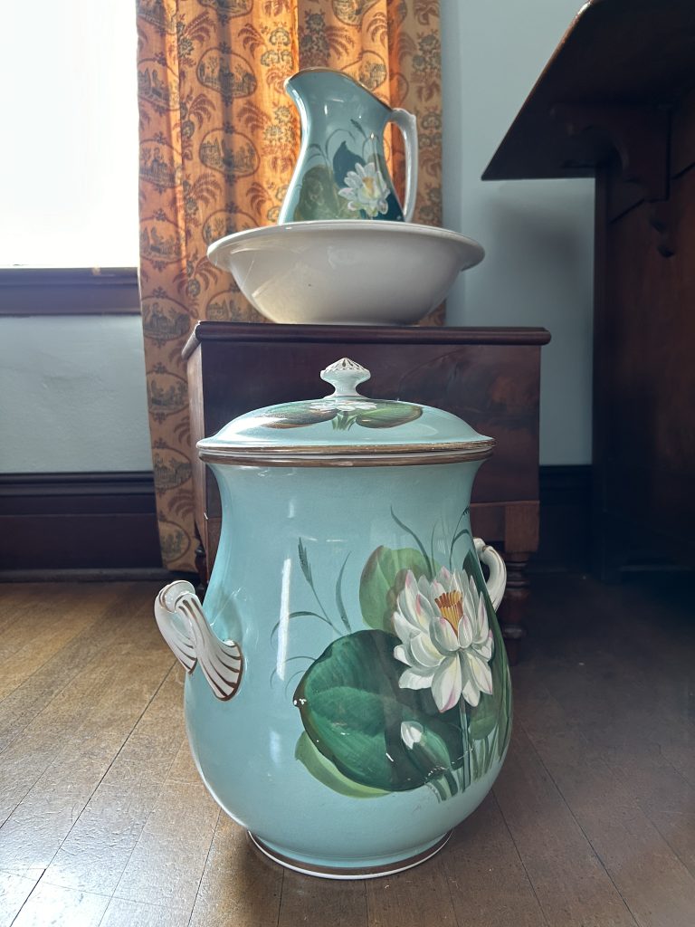 An image of a large blue pot with a lid and white handles. On the front there is an image of a white water lily. Behind there is a box and another pitcher with lily's sitting in a bowl.