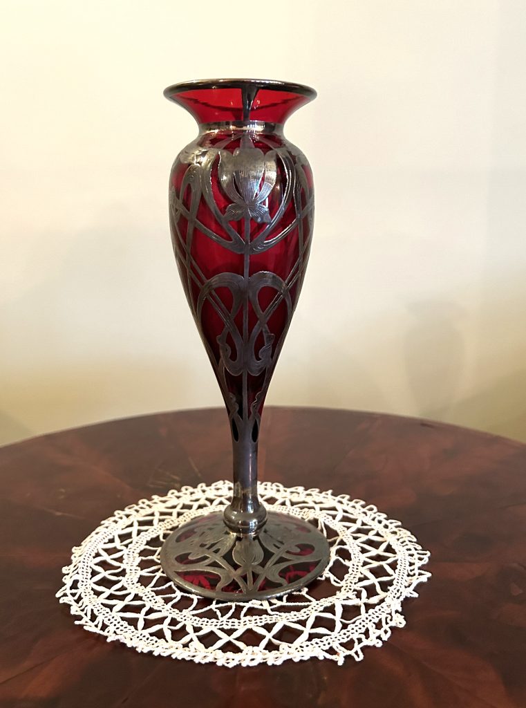 A red vase that has a round opening and then a thin neck. The vase tapers to the bottom where it has a round foot. The glass is red, though there is silver overlay all over. The piece sits on a white doily on a wood table.