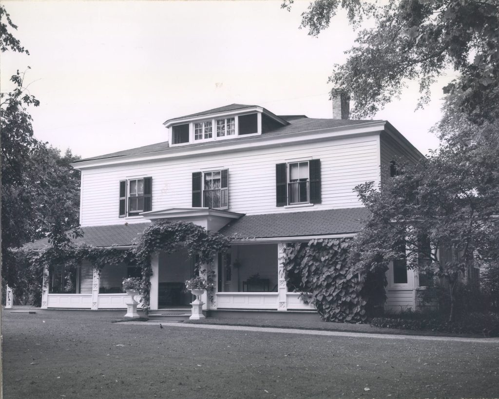 A black and white image of a house with dark shutters. The house is covered with grapevines.