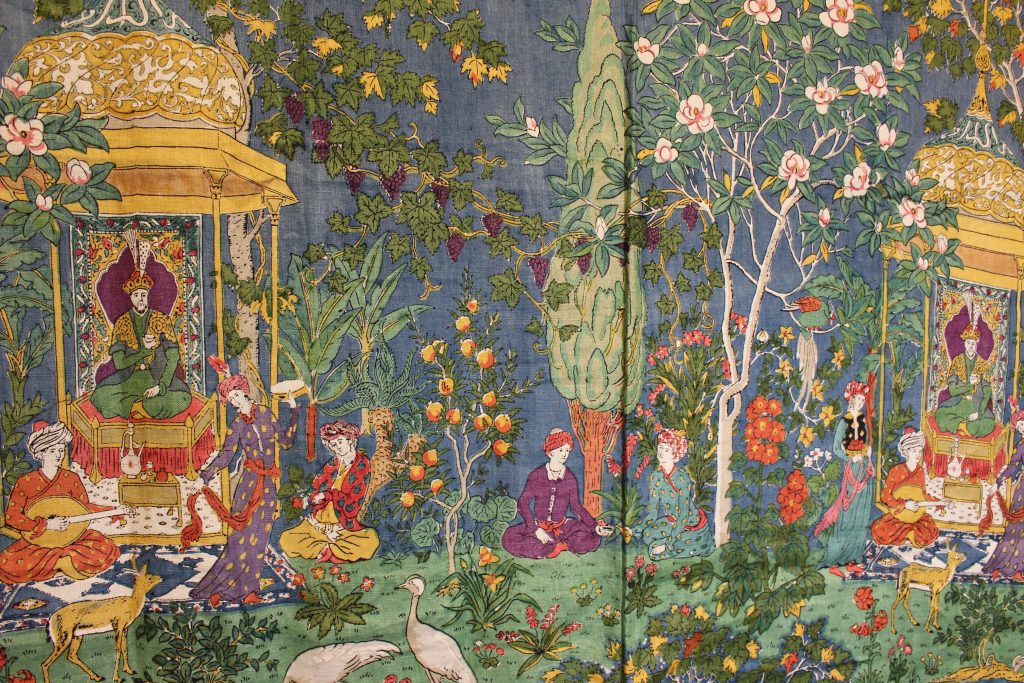 A large blue tapestry with image of a person in a pagoda and another group sitting playing a sitar. They are surrounded by foliage.
