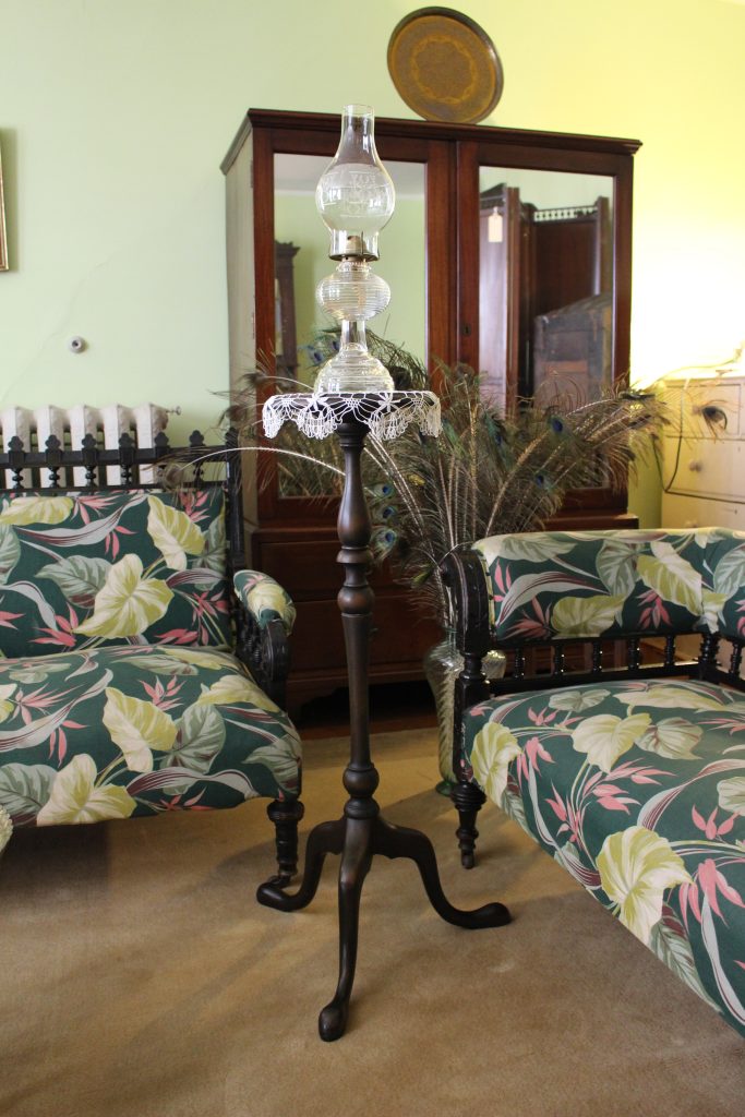 An image of a tall skinny table with a glass lamp on top. It is sitting in a room with tropical patterned green sofas.