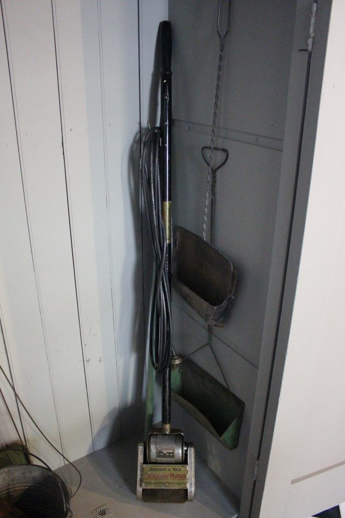 An image of a grey closet. Inside there is a floor polisher, which looks like a thin vacuum.