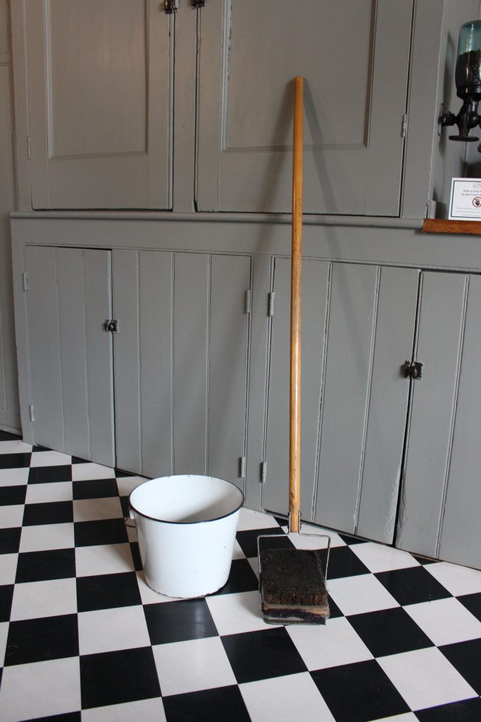 An image of a mop like item with a metal head. It is standing next to a bucket on a black and white checkered floor with grey cabinets in the background.