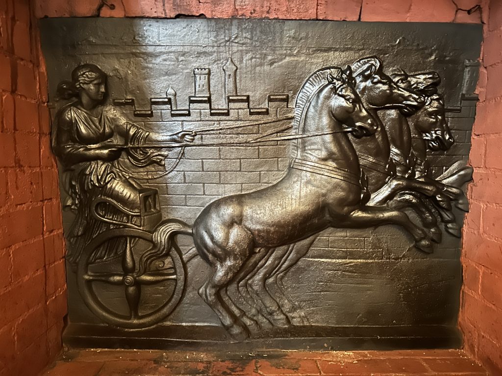 A red brick area. In the center there is an iron picture of a woman in a chariot. She is holding reins driving four horses.
