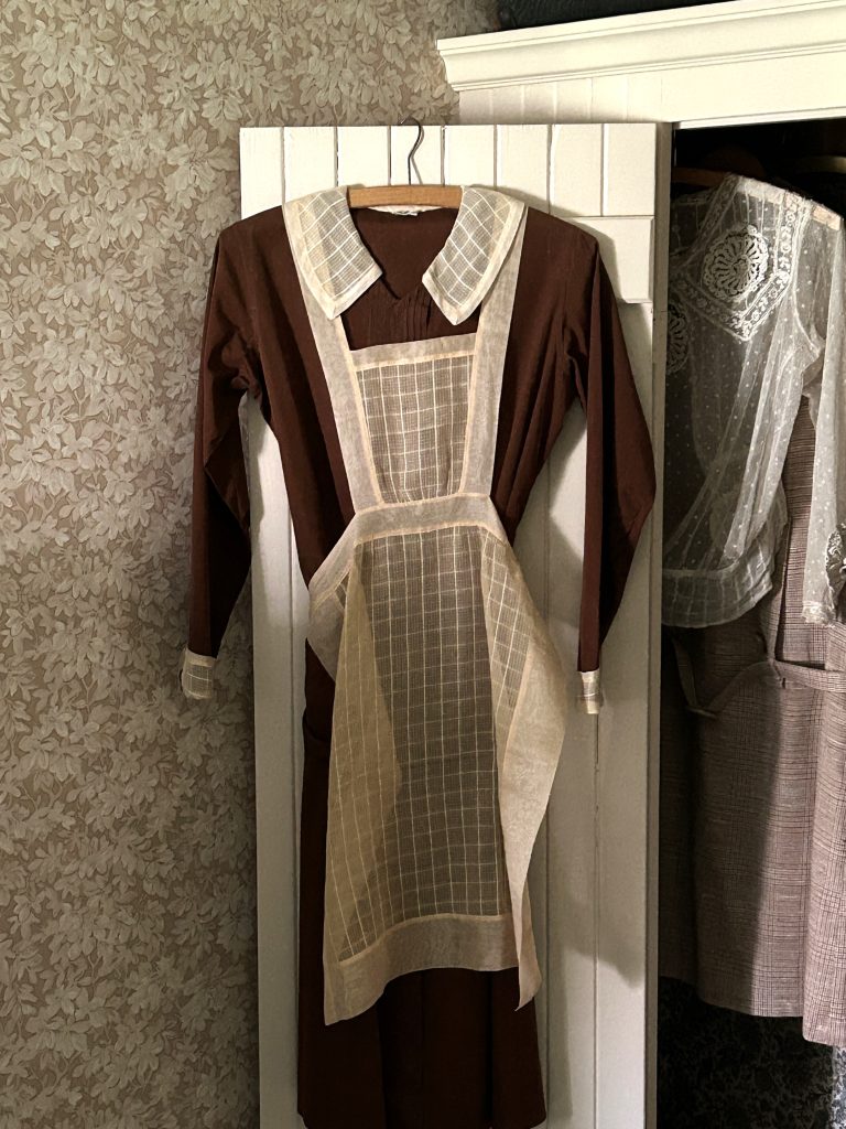 A brown dress handing on a white closet door. Overtop there is a cream apron that is sheer. The same material matches the cuffs and collar of the dress.