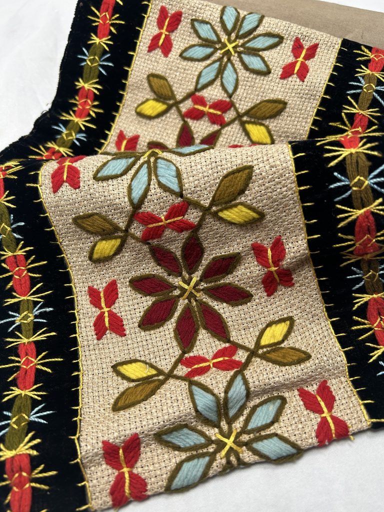 An image of fabric that has been crinkled. On it, the center is beige with geometric flowers and leaves in blue, yellow, red, and green. it has black borders on either side and some orange and green on it.