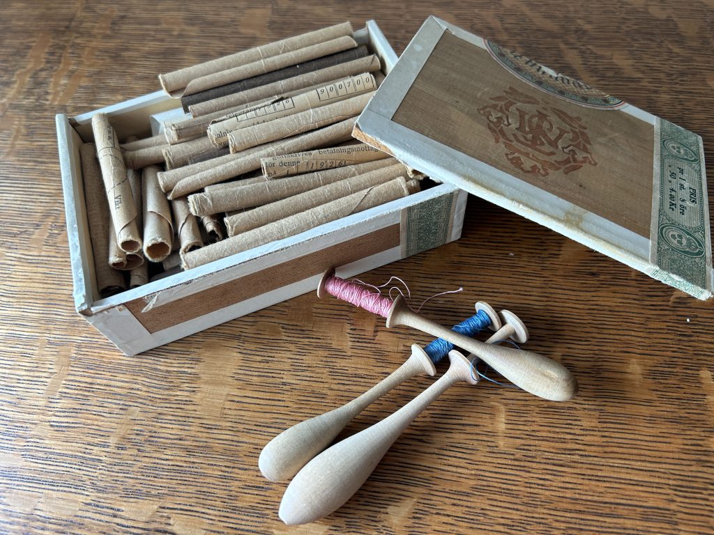 A wooden box with the lid partially off. Inside there are rolled up cigar papers. Outside there are three bobbins, one with pink thread, one with blue, and one with none.