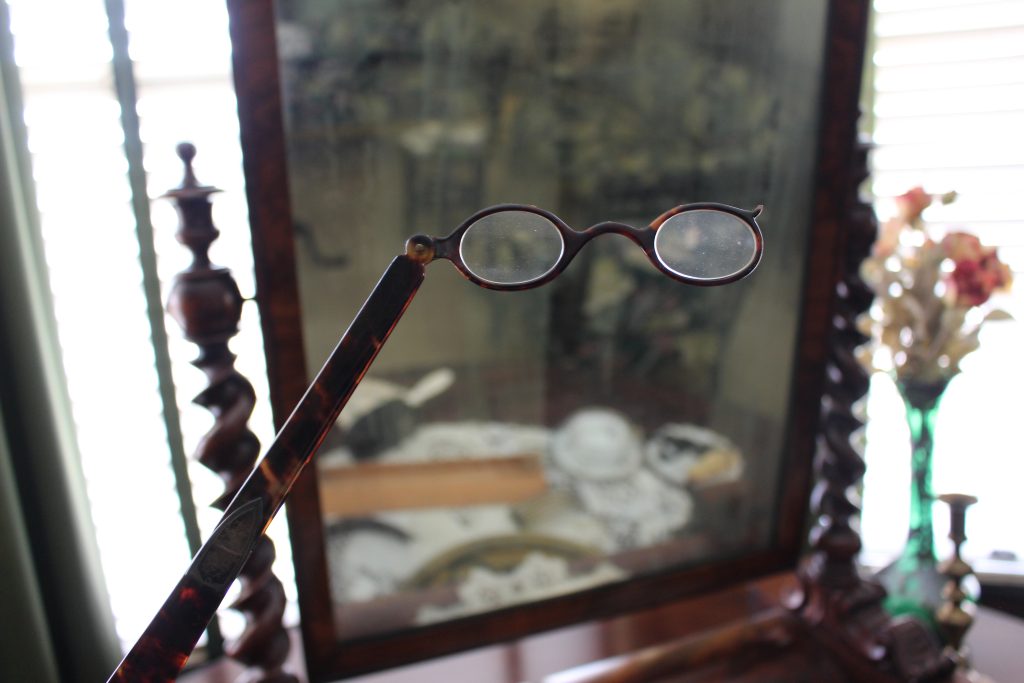 An image of a pair of small glasses with tortoise shell rims. On the left side there is a long handle being held up. In the background, there is a mirror and other beauty items on a table.