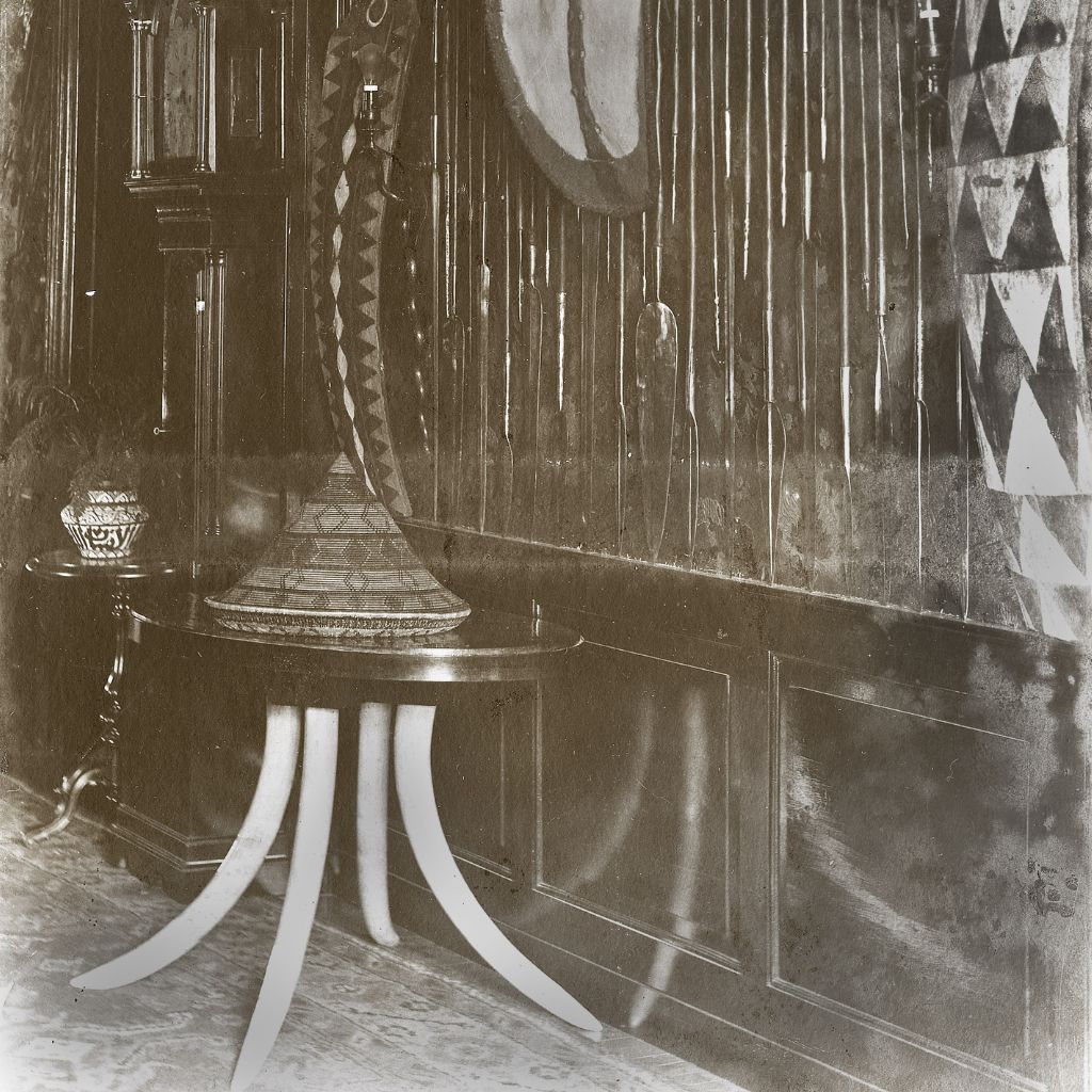 A historical image of a hallway. In the forefront there is a table with curved elephant tusks. On the wall are decorated with patterned shields.