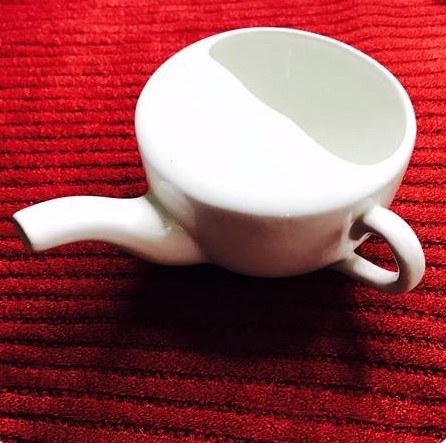 A small white teacup item with a small handle. It is sitting on a red pillow.