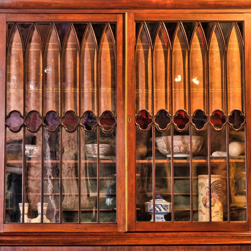 A wooden cabinet with glass doors. On the door there are arched patterns which frame the spines of books. Below shelves can be seen with pottery on them.