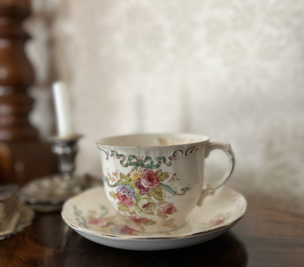 An image of a large white teacup with purple and green roses and a blue bow. It is sitting on a wood table.