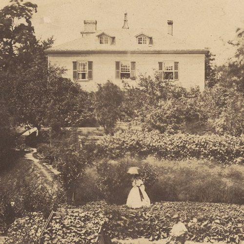 A sepia image of a white house. There are extensive gardens in the front and there is a lady with a parasol on the lawn as well as a man gardening.