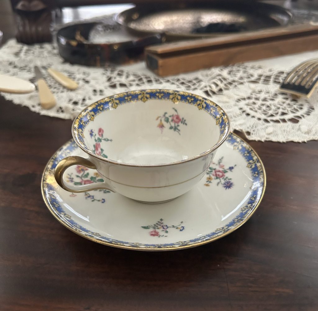 An image of a white teacup with a blue and gold border on the top edge and on the saucer. Inside the cup and around the edge of the saucer is pink roses with vines. It is on a wooden table with hair and makeup items in the background.